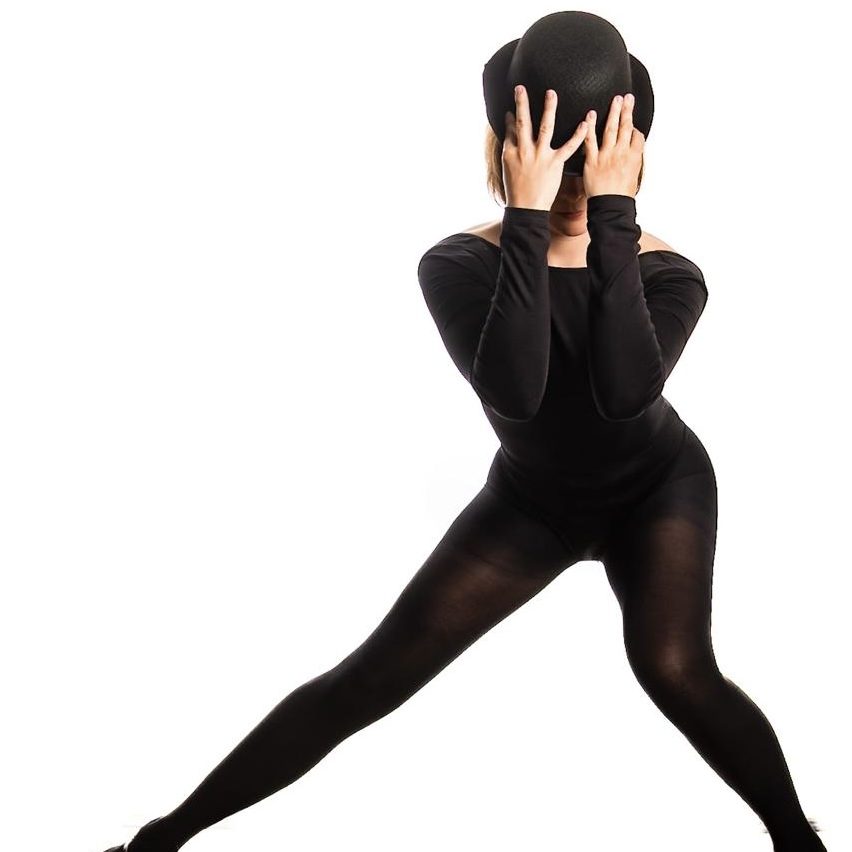 Dance in black leotard, black tights and black heels wearing a black bowler hat covering her face with her hands.